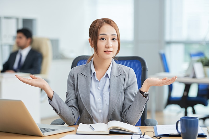 asian woman sitting desk office looking camera with helpless hand gesture